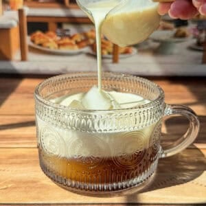 A hand pours creamy milk from a small glass pitcher into a patterned glass mug filled with coffee, set on a wooden surface with sunlight streaming in.