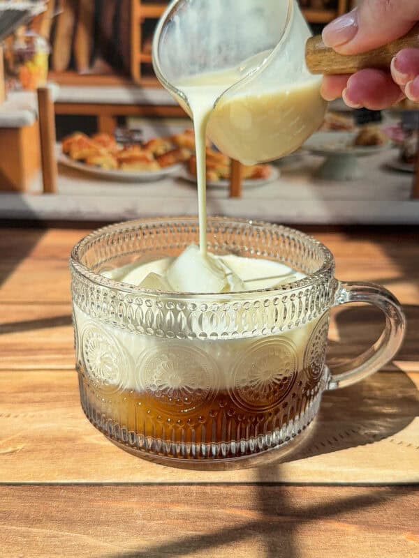 A hand pours creamy milk from a small glass pitcher into a patterned glass mug filled with coffee, set on a wooden surface with sunlight streaming in.