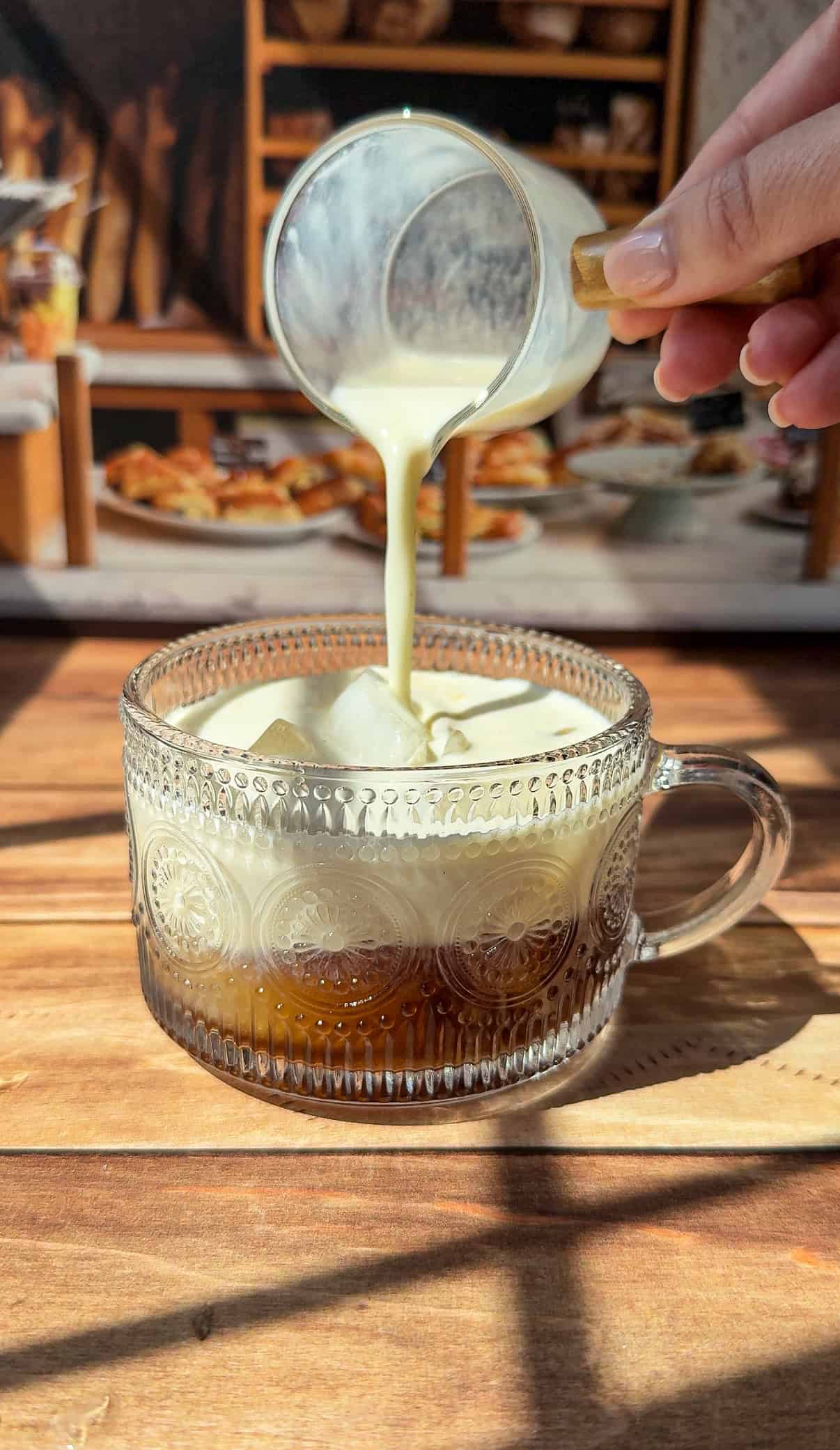 A hand pours creamy banana milk latte into a glass mug of iced coffee, sitting on a wooden table with a cozy kitchen background in soft sunlight.