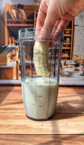 A hand holding half a peeled banana is about to place it into a blender cup partially filled with milk, on a wooden surface with a kitchen in the background.