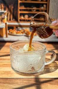 A hand pours espresso from a small glass pitcher over a scoop of vanilla ice cream in a decorative glass cup, creating an affogato. The scene is sunlit with baked goods and shelves in the blurred background.