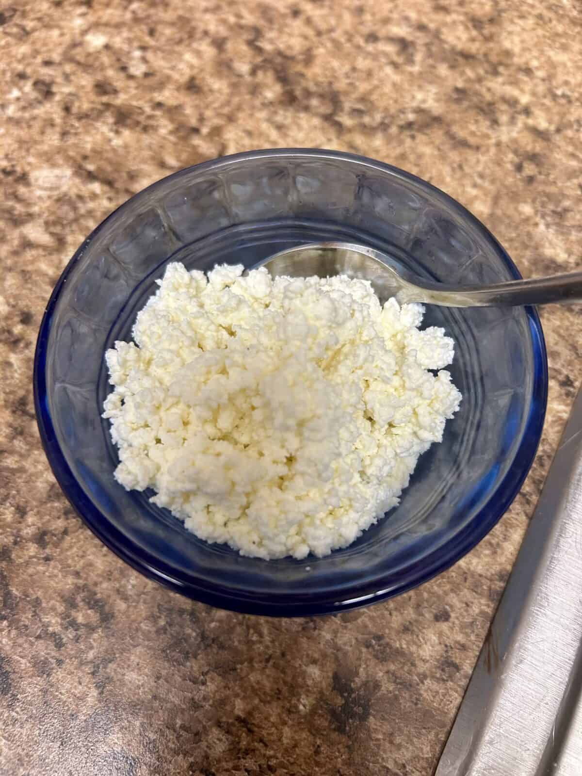 A small blue glass bowl filled with white, crumbly cottage cheese, with a metal spoon resting inside, placed on a brown, speckled countertop.