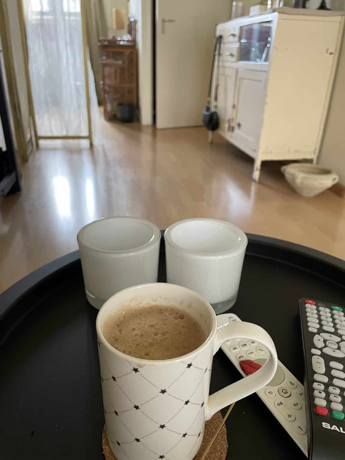 A patterned mug of Banana Milk Latte sits on a black tray with two white candles and several remote controls in a cozy, sunlit living room with wooden floors and light-colored furniture.