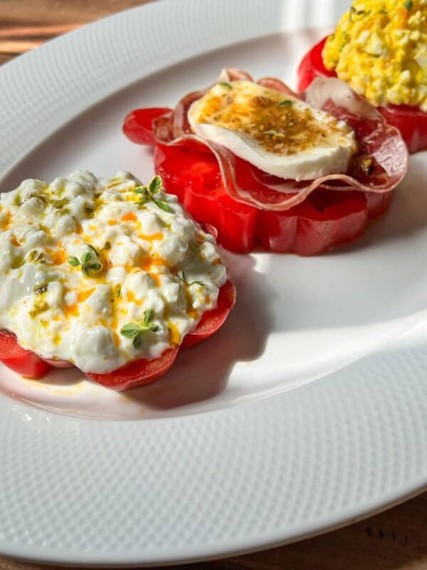 Three tomato slices topped with different ingredients sit on a white plate: one with cottage cheese and herbs, one with prosciutto and soft cheese, and one with an egg salad mixture.