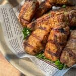 A bowl lined with newspaper holds several grilled, golden-brown sausages or meat rolls, garnished with fresh parsley and bathed in warm, natural sunlight.
