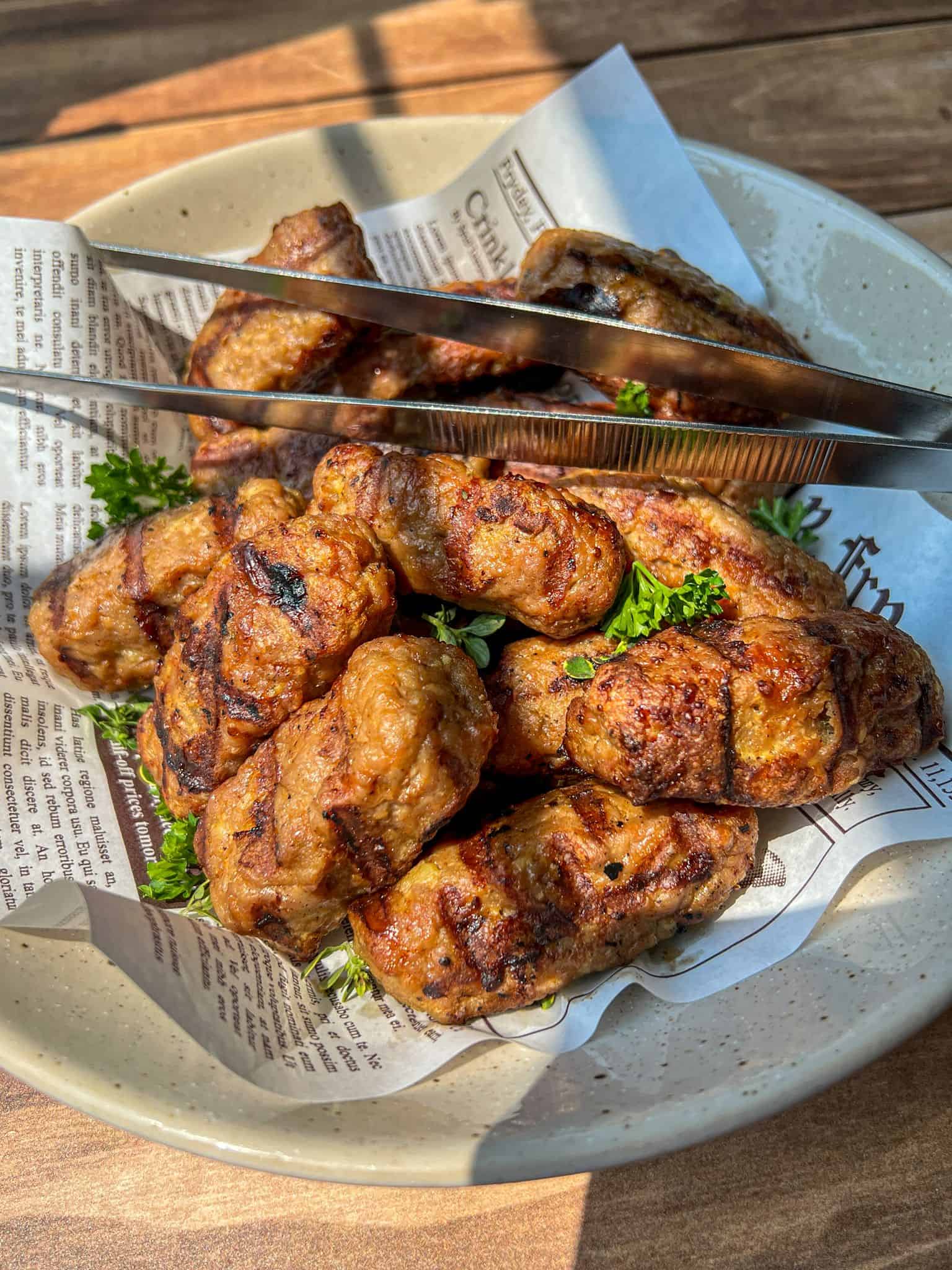 A plate of grilled meat sausages, or Turkey Cevapi, garnished with parsley, served on newspaper-style paper, with metal tongs resting on top. Sunlight shines onto the dish, highlighting the texture of the sausages.
