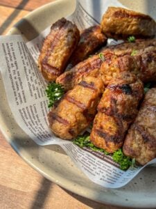 A bowl lined with newspaper holds several grilled, golden-brown sausages or meat rolls, garnished with fresh parsley and bathed in warm, natural sunlight.