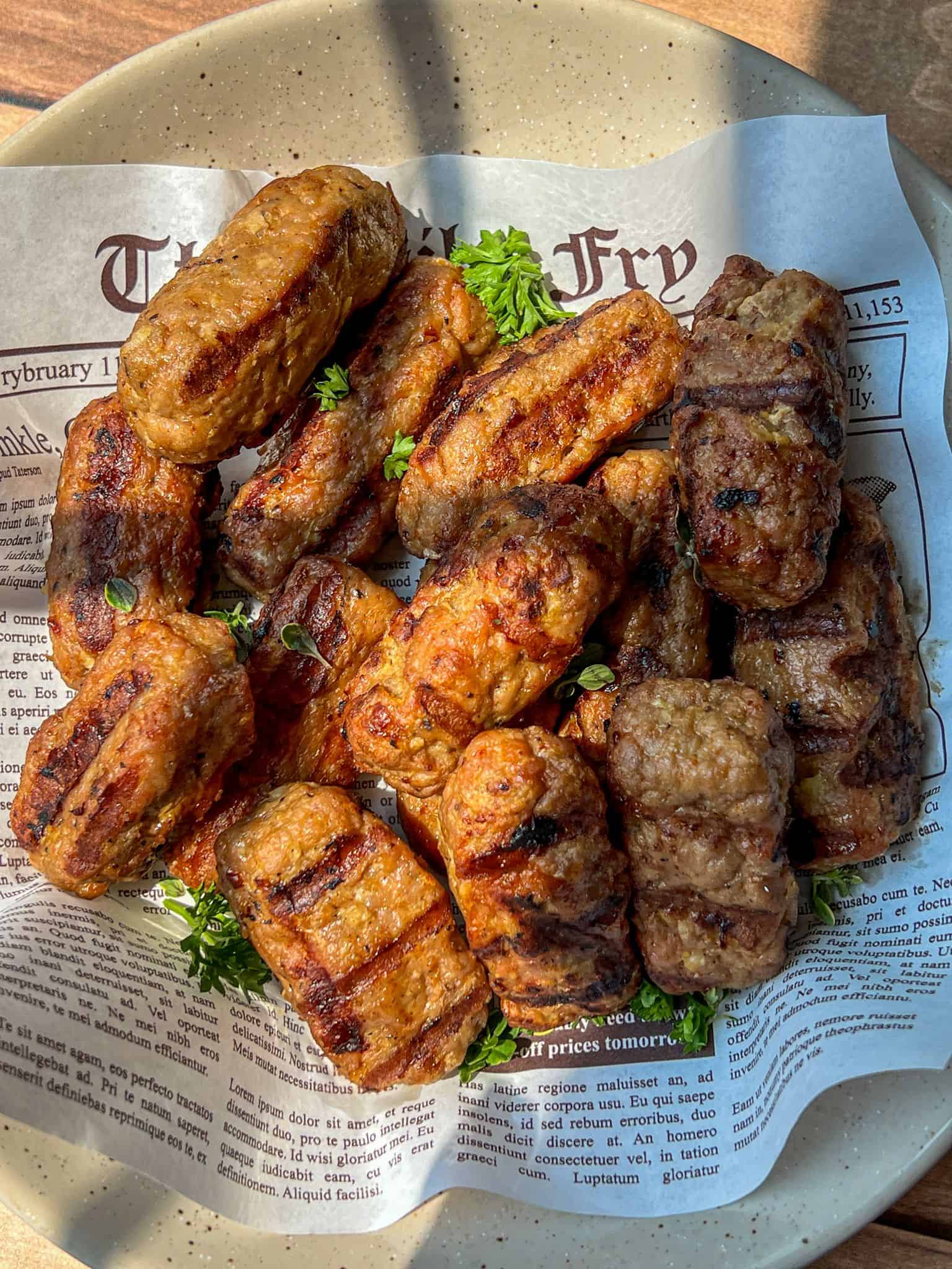 Grilled beef and pork sausages, known as Turkey Cevapi are served on a plate lined with newspaper-style paper and garnished with fresh parsley. The sausages are browned with visible grill marks.