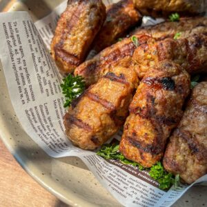 A bowl lined with newspaper holds several grilled, golden-brown sausages or meat rolls, garnished with fresh parsley and bathed in warm, natural sunlight.