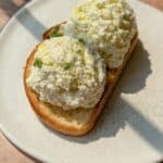 A slice of toasted bread topped with two generous scoops of ricotta cheese, garnished with lemon zest and fresh herbs, served on a white plate.