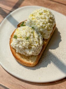 A slice of toasted bread topped with two generous scoops of ricotta cheese, garnished with lemon zest and fresh herbs, served on a white plate.