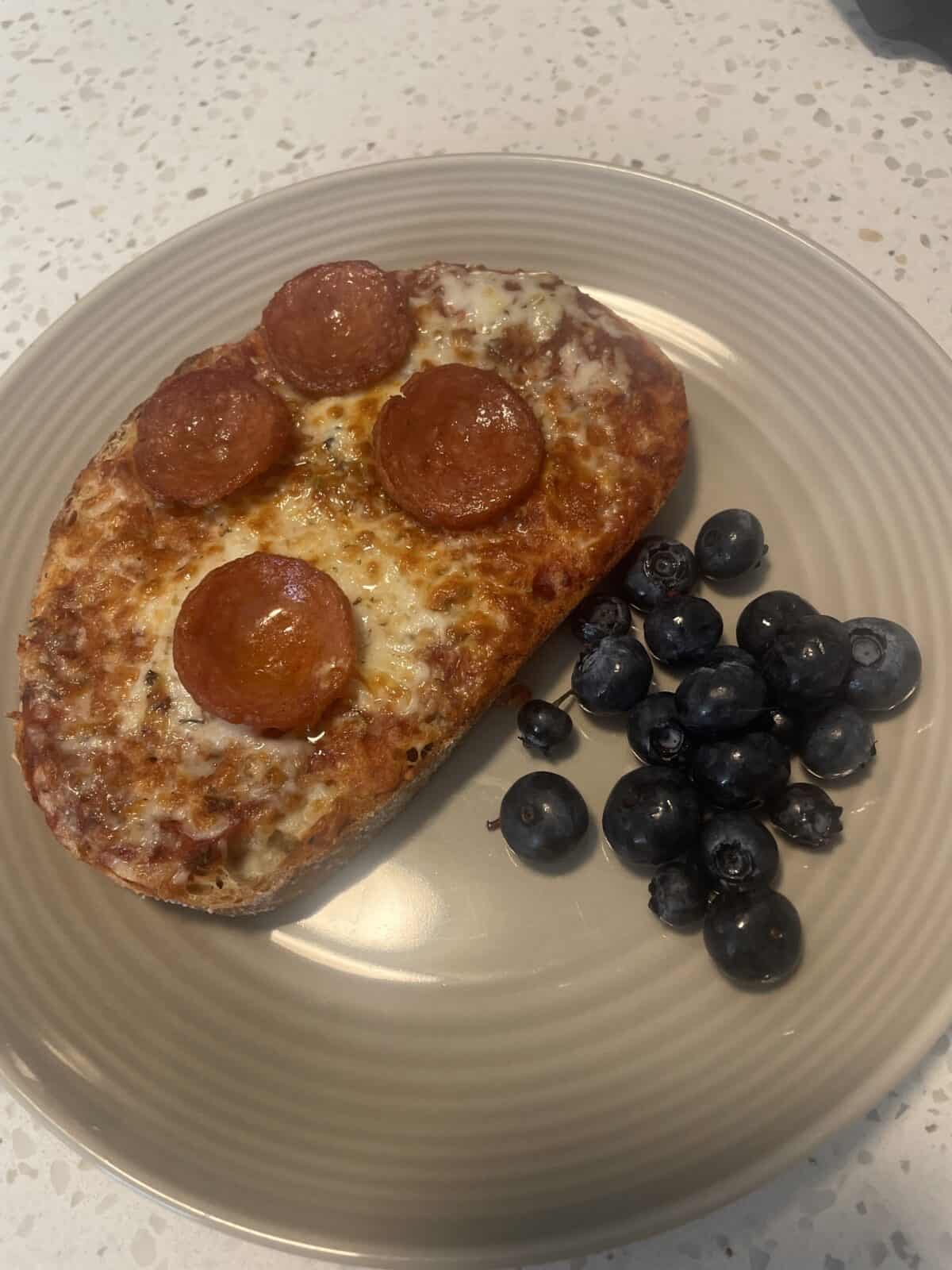 A plate with a slice of pepperoni pizza toast topped with four pepperoni slices, next to a small pile of fresh blueberries on a beige plate.