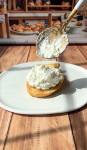 A spoon spreads a generous dollop of clotted cream onto a halved scone on a white plate, with a bakery display featuring pastries in the blurred background.