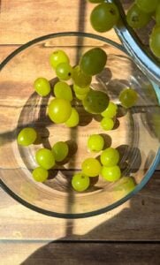 Green grapes are being poured into a clear glass bowl on a wooden surface, with sunlight casting shadows across the scene.