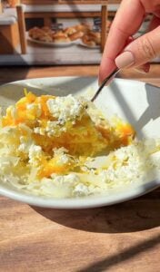 A hand holding a fork is mixing shredded cheese and crumbled white cheese in a white bowl on a wooden table, with a bakery display blurred in the background.