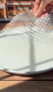 A hand places a sheet of translucent rice paper onto a white plate, with sunlight casting shadows across the scene and baked goods visible in the background.