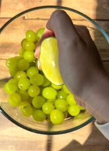 A hand squeezes a halved lime over a glass bowl filled with green grapes on a wooden surface. Sunlight casts shadows across the scene.