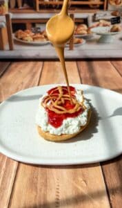 A spoon drizzles peanut butter over a biscuit topped with cottage cheese and strawberry jam, set on a white plate on a wooden table.