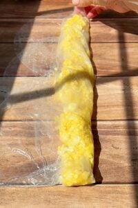 A hand rolls a translucent rice paper sheet filled with scrambled eggs on a wooden surface, in bright sunlight with visible shadows.