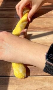 Hands are twisting a long piece of yellow dough on a wooden surface. One hand wears a smartwatch with a black band. Sunlight casts shadows across the scene.