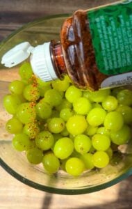 A bottle of chili powder is being sprinkled over a glass bowl filled with fresh green grapes on a wooden surface.