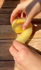 A person shapes a piece of yellow dough on a wooden surface, using both hands. Sunlight casts shadows on the table and hands.