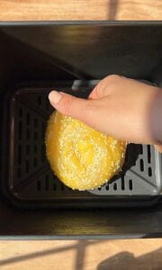 A hand places a round, sesame-seeded dough into a black air fryer basket, ready for baking. The surface beneath the air fryer is wooden, and sunlight casts soft shadows over the scene.