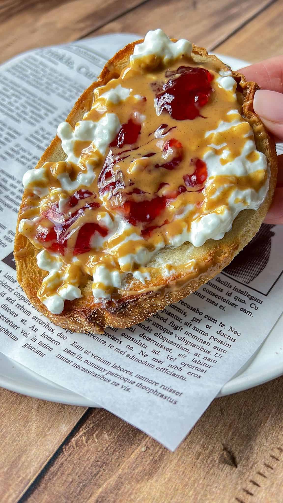 A hand holds a slice of toasted bread topped with peanut butter, dollops of cottage cheese, and swirls of red jelly, resting on a plate lined with printed newspaper-style parchment.