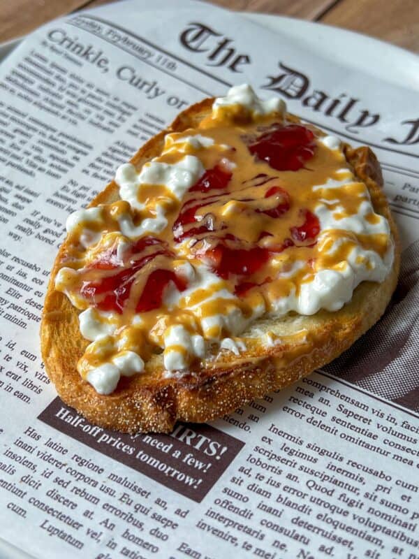 A slice of toasted bread topped with cottage cheese, strawberry jam, and drizzled peanut butter sits on a plate designed to look like a newspaper page.