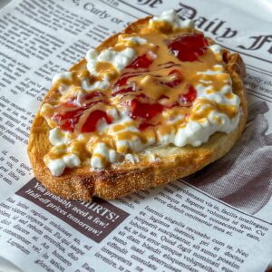 A slice of toast topped with cottage cheese, peanut butter, and strawberry jelly sits on a plate designed to look like a newspaper page.