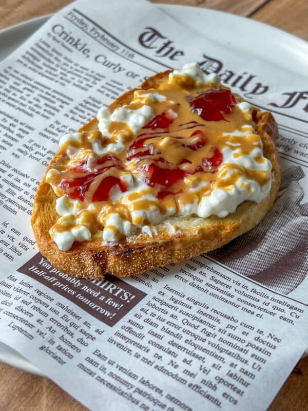 A slice of toast topped with cottage cheese, peanut butter, and strawberry jelly sits on a plate designed to look like a newspaper page.