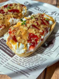 Two slices of bread topped with creamy cheese, red jam, a drizzle of caramel or honey, crunchy crumbs, and fresh herbs, served on a plate lined with newspaper-style paper.