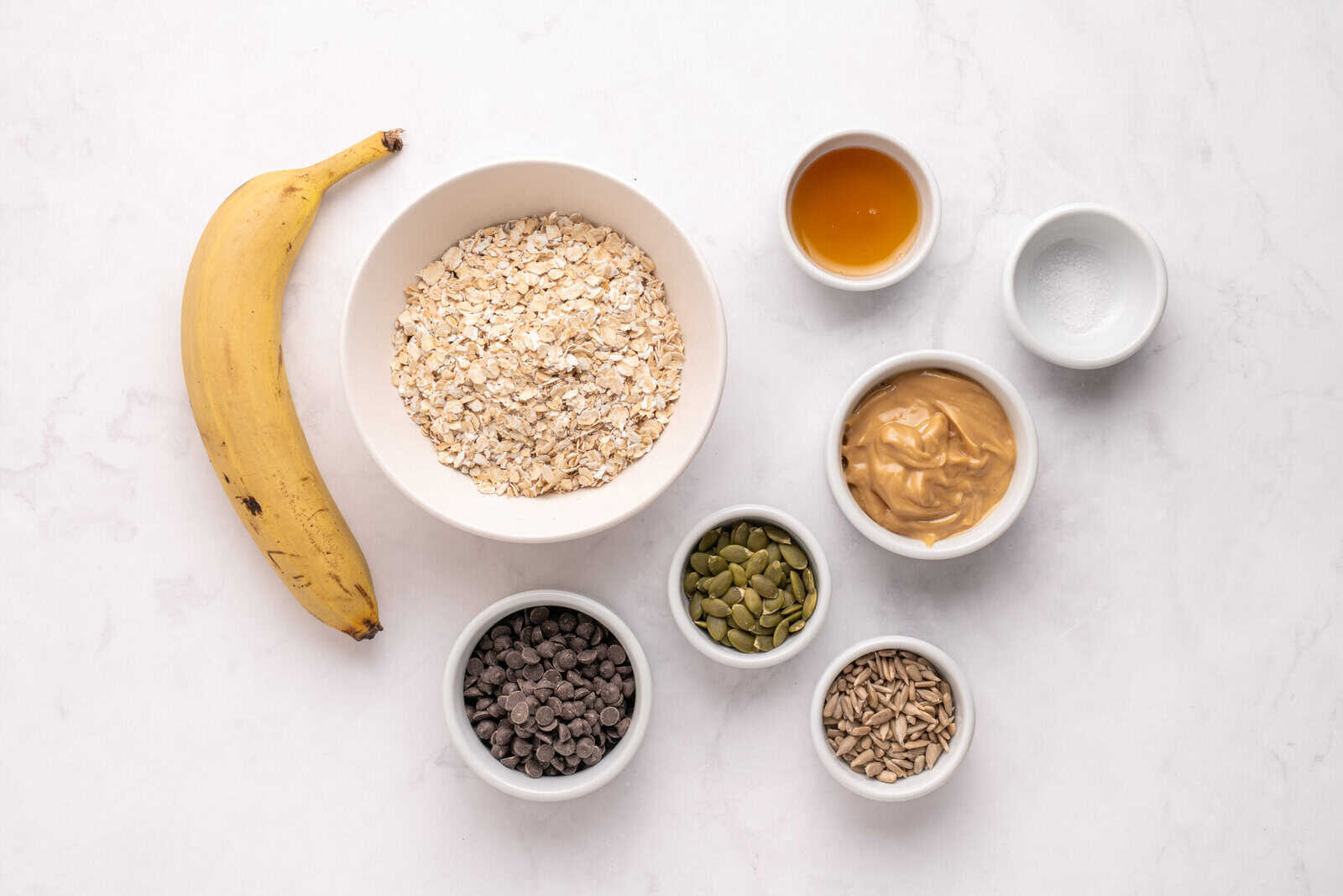 A banana, a bowl of rolled oats, and small bowls of honey, peanut butter, salt, pumpkin seeds, chocolate chips, and sunflower seeds are arranged on a white marble surface.