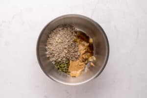 A metal bowl containing rolled oats, sunflower seeds, pumpkin seeds, peanut butter, honey, and mashed banana on a light marble surface.
