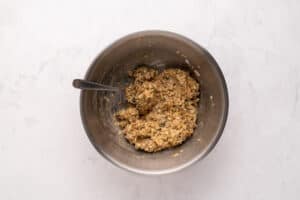 A metal mixing bowl with a spoon inside, containing a thick, mixed batter or dough with visible grains and herbs, placed on a light marble surface.