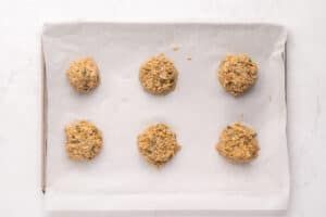 Six scoops of unbaked cookie dough spaced evenly on a parchment-lined baking sheet, ready to be baked, set on a white marble surface.