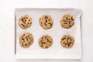 Six unbaked chocolate chip cookies are arranged on a baking sheet lined with parchment paper, ready to be baked. The cookies are spaced evenly apart on a white surface.