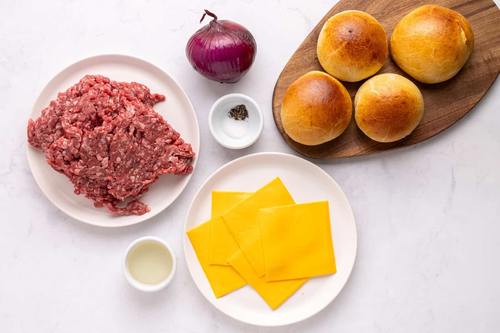 Top-down view of burger ingredients: a plate of ground beef, a red onion, salt and pepper, four burger buns on a wooden board, American cheese slices on a plate, and a small bowl of oil on a white surface.