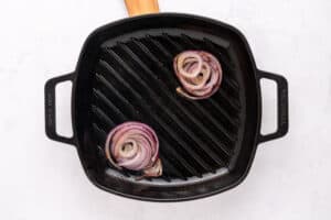 A black cast iron grill pan with two slices of red onion being grilled, one near the top right and one near the bottom left, on a white surface.