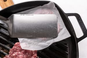 A metal spatula presses ground meat on parchment paper in a cast iron grill pan, partially flattening the meat for cooking.