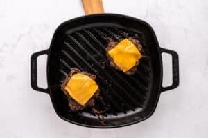 A black grill pan with two cooked hamburger patties, each topped with a slice of melted cheddar cheese, sits on a light-colored surface. The handle of the grill pan is wooden.