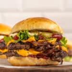 A double cheeseburger with two beef patties, cheddar cheese, lettuce, pickles, ketchup, mayonnaise, and grilled onions in a brioche bun, served on a wooden board.