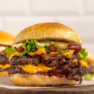 A double cheeseburger with two beef patties, cheddar cheese, lettuce, pickles, ketchup, mayonnaise, and grilled onions in a brioche bun, served on a wooden board.