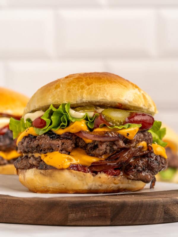 A double cheeseburger with two beef patties, cheddar cheese, lettuce, pickles, ketchup, mayonnaise, and grilled onions in a brioche bun, served on a wooden board.