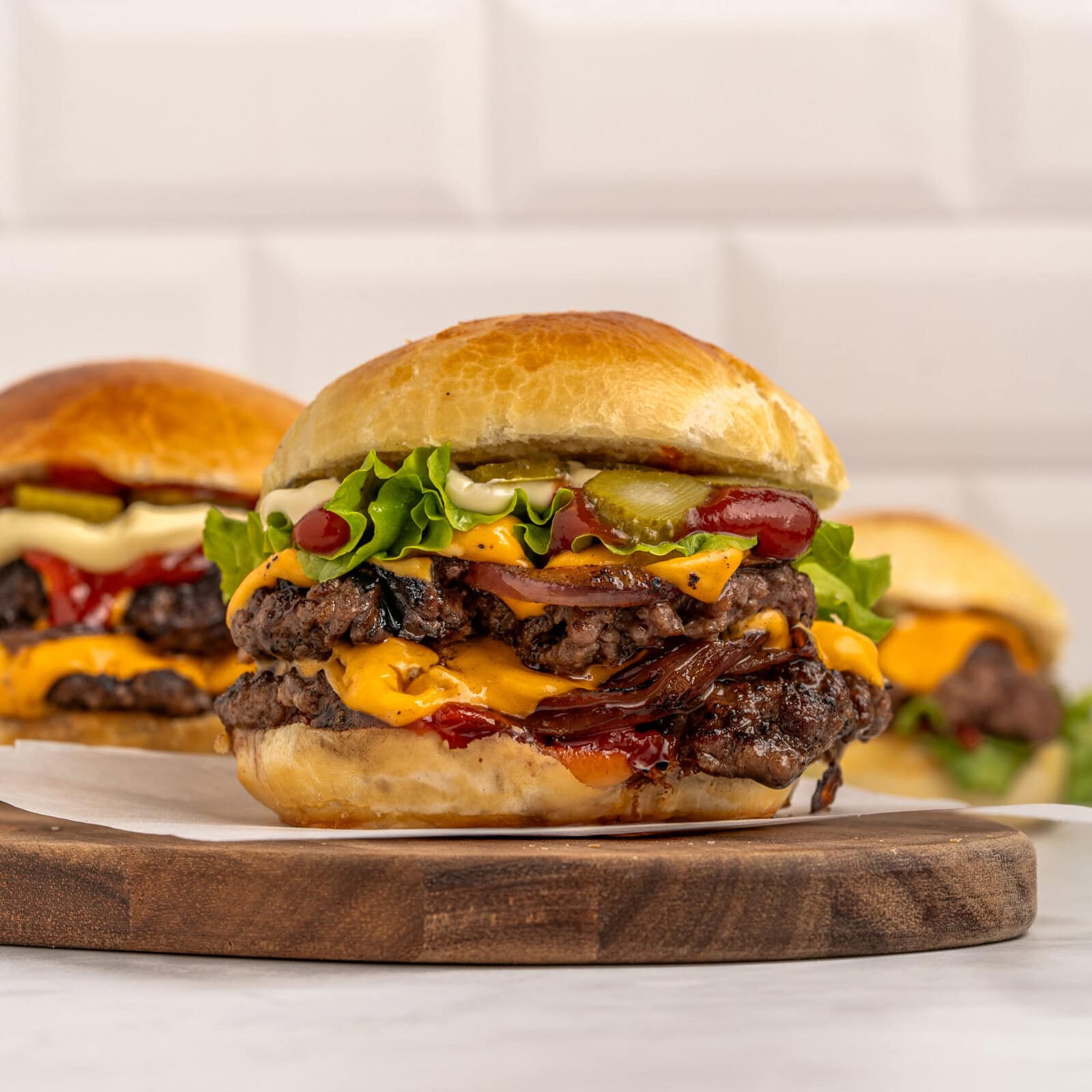 A double cheeseburger with lettuce, tomato, pickles, onions, and melted cheese on a brioche bun, placed on a wooden board, with two more burgers blurred in the background.