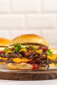 A juicy double cheeseburger with melted cheese, crispy bacon, lettuce, pickles, ketchup, and mayo on a toasted bun, sitting on a wooden board with another burger in the background.