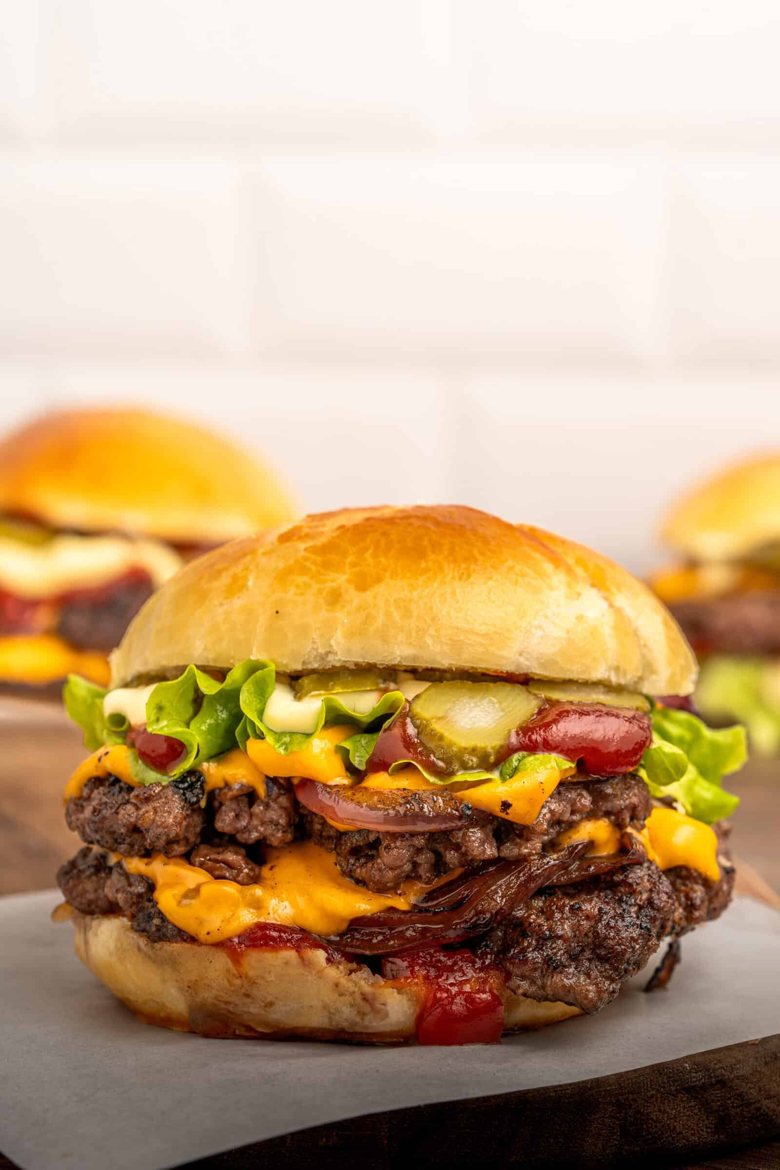 A close-up of a cheeseburger with two beef patties, melted cheddar cheese, lettuce, pickles, onions, ketchup, and mayonnaise in a shiny brioche bun. Two more burgers are blurred in the background.
