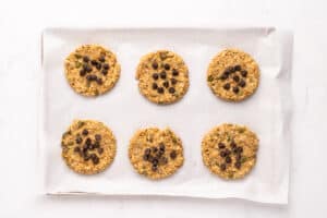 Six round oatmeal cookies are arranged in two rows on a parchment-lined baking sheet. Some cookies are topped with chocolate chips, while others have seeds. The background is a white, marble-like surface.