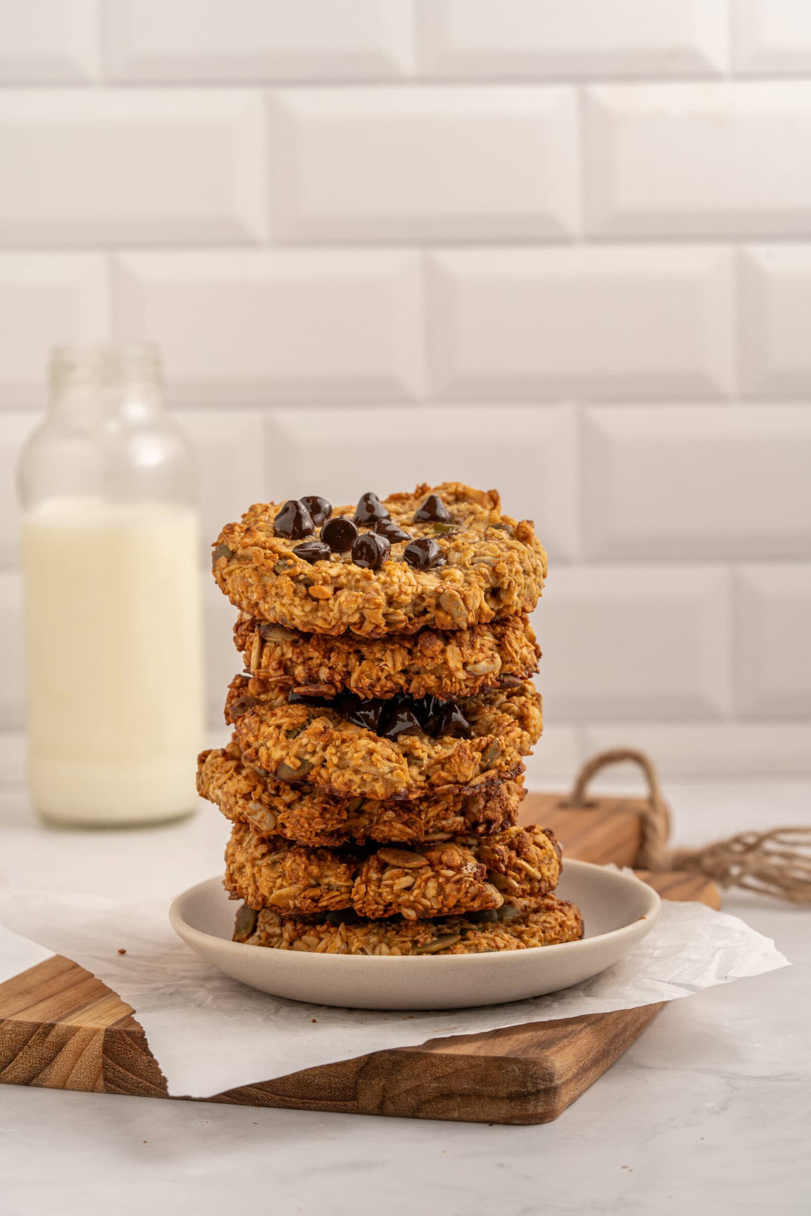 A stack of chocolate chip cookies sits on a white plate, with a bottle of milk in the background. The scene is set against a white tiled wall on a light countertop.