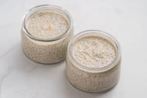 Two small glass jars filled with creamy chia seed pudding are placed on a white marble surface. The pudding is light beige with visible chia seeds evenly dispersed throughout.
